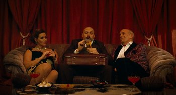Movie still from “Bronson” (2008), directed by Nicolas Winding Refn – A man sitting on a couch next to two other people; Wide shot, Low angle