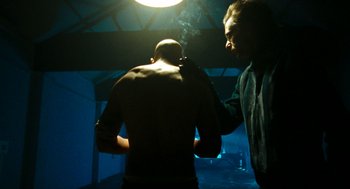 Movie still from “Bronson” (2008), directed by Nicolas Winding Refn – A man standing next to another man in a dark room; Medium shot, Over the shoulder angle