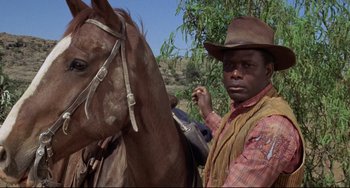 Movie still from “Buck and the Preacher” (1972), directed by Joseph Sargent – A man standing next to a brown horse on a field; Close Up shot, Low angle