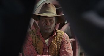Movie still from “Buck and the Preacher” (1972), directed by Joseph Sargent – A man wearing a cowboy hat sitting in front of a wheel; Close Up shot, Over the shoulder angle