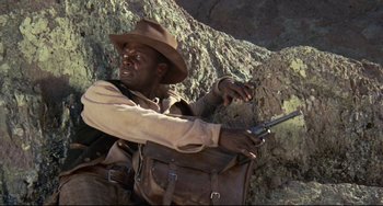 Movie still from “Buck and the Preacher” (1972), directed by Joseph Sargent – A man holding a gun while standing next to a rock wall; Medium shot, Low angle