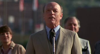Movie still from “Bull Durham” (1988), directed by Ron Shelton – An older man wearing a suit and tie while standing in front of a microphone; Close Up shot, Low angle