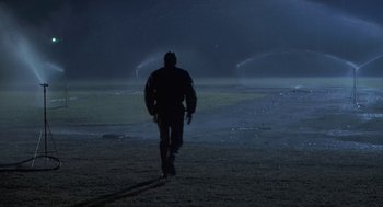 Movie still from “Bull Durham” (1988), directed by Ron Shelton – A man walking in a field at night with water sprinklers in the background; Wide shot, Over the shoulder angle