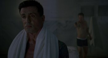 Movie still from “Bullet to the Head” (2012), directed by Walter Hill – A man with a towel around his neck is looking at the camera while another man takes a picture in the background; Medium shot, Over the shoulder angle