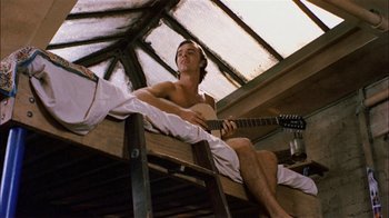 Movie still from “Butterflies Are Free” (1972), directed by Milton Katselas – A shirtless man sitting on top of a ladder holding a guitar; Medium shot, Overhead angle