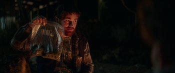 Movie still from “Cabin Fever” (2016), directed by Travis Zariwny – A man sitting in the dark holding a bag; Medium shot, Over the shoulder angle
