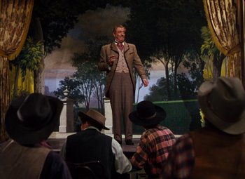 Movie still from “Calamity Jane” (1953), directed by David Butler – A man standing on a stage in front of a group of people; Wide shot, Low angle