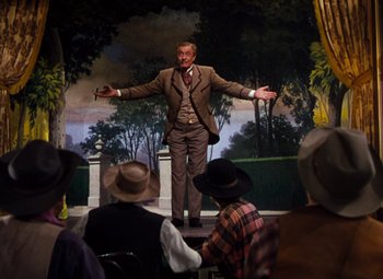 Movie still from “Calamity Jane” (1953), directed by David Butler – A man in a brown suit is on a stage with a microphone; Wide shot, Low angle