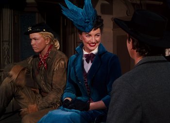 Movie still from “Calamity Jane” (1953), directed by David Butler – A woman in a blue suit and hat sitting next to two men; Medium shot, Over the shoulder angle