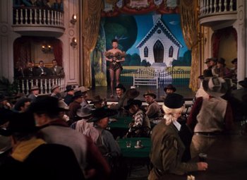 Movie still from “Calamity Jane” (1953), directed by David Butler – A group of people sitting in a room with a painting of a church; Wide shot, Low angle