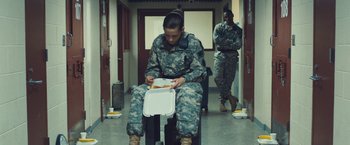 Movie still from “Camp X-Ray” (2014), directed by Peter Sattler – A woman in fatigues sitting on a chair eating food; Wide shot, High angle