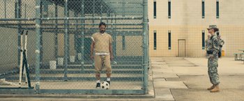 Movie still from “Camp X-Ray” (2014), directed by Peter Sattler – A man standing in front of a fence holding a soccer ball; Wide shot, Low angle