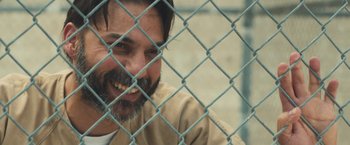 Movie still from “Camp X-Ray” (2014), directed by Peter Sattler – A man with a goatee is smiling behind a chain link fence; Close Up shot, Over the shoulder angle