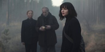 Movie still from “Dark” (2017), created by Baran bo Odar – A woman standing next to a man and a woman holding an apple; Medium shot, Over the shoulder angle