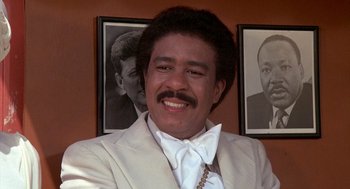 Movie still from “Car Wash” (1976), directed by Michael Schultz – A man in a white suit and a white bow tie in front of a picture of himself; Close Up shot, Low angle
