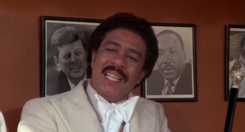 Movie still from “Car Wash” (1976), directed by Michael Schultz – A man in a suit and tie next to a picture of john f; Close Up shot, Low angle
