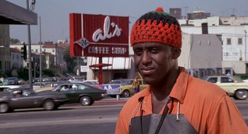 Movie still from “Car Wash” (1976), directed by Michael Schultz – A man wearing an orange shirt and a crocheted hat stands on the side of the road; Close Up shot, Low angle
