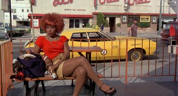 Movie still from “Car Wash” (1976), directed by Michael Schultz – A woman sitting on top of a bench next to a yellow taxi cab; Wide shot, Low angle
