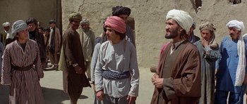 Movie still from “Caravans” (1978), directed by James Fargo – A group of men standing next to each other on a dirt road; Medium shot, Over the shoulder angle