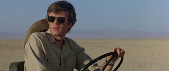 Movie still from “Caravans” (1978), directed by James Fargo – A man driving a car on a dirt road in the desert; Close Up shot, Low angle
