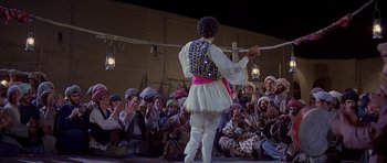 Movie still from “Caravans” (1978), directed by James Fargo – A group of people sitting and standing around a man on a stage; Wide shot, High angle
