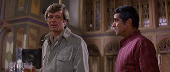 Movie still from “Caravans” (1978), directed by James Fargo – Two men standing next to each other in front of a building; Medium shot, Over the shoulder angle