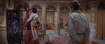 Movie still from “Caravans” (1978), directed by James Fargo – A group of people standing in a living room; Wide shot, Over the shoulder angle