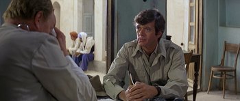 Movie still from “Caravans” (1978), directed by James Fargo – A man sitting at a table with a pen in his mouth; Close Up shot, Over the shoulder angle