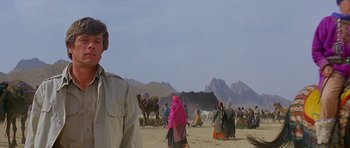 Movie still from “Caravans” (1978), directed by James Fargo – A group of people standing on top of a sandy beach; Medium shot, Over the shoulder angle