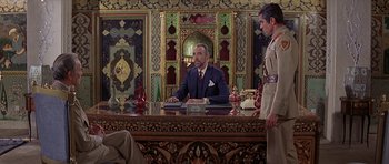 Movie still from “Caravans” (1978), directed by James Fargo – A man sitting at a desk in front of two other men; Wide shot, Over the shoulder angle