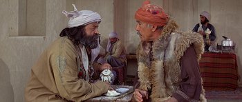 Movie still from “Caravans” (1978), directed by James Fargo – Two men are sitting at a table and talking to each other; Medium shot, Over the shoulder angle