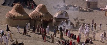 Movie still from “Caravans” (1978), directed by James Fargo – A group of people standing around a tent on a dirt ground; Extreme Wide shot, High angle