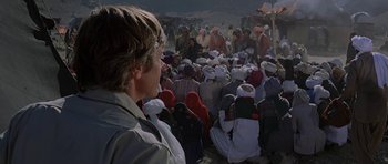 Movie still from “Caravans” (1978), directed by James Fargo – A crowd of people sitting on the ground in a field; Medium shot, Over the shoulder angle