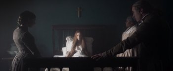 Movie still from “Carmilla” (2019), directed by Emily Harris – A woman sitting on a bed in a dark room next to a cross; Medium shot, Over the shoulder angle