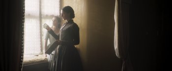 Movie still from “Carmilla” (2019), directed by Emily Harris – A woman in a black dress standing next to a window; Medium shot, Over the shoulder angle