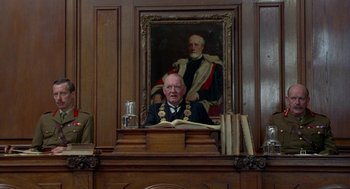 Movie still from “Carrington” (1995), directed by Christopher Hampton – A man is sitting at a podium in front of a painting; Wide shot, Low angle