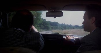 Movie still from “Castle Freak” (1995), directed by Stuart Gordon – A man driving a car on a road with trees in the background; Medium shot, Over the shoulder angle