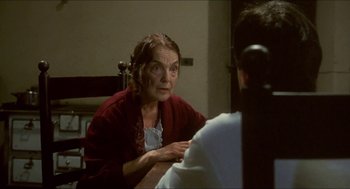 Movie still from “Castle Freak” (1995), directed by Stuart Gordon – An older woman sitting in front of a man in a room; Medium shot, Over the shoulder angle