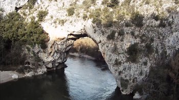 Movie still from “Cave of Forgotten Dreams” (2010), directed by Werner Herzog – A river flowing through a rock arch in the middle of a forest; Extreme Wide shot, High angle
