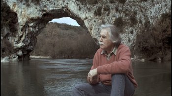 Movie still from “Cave of Forgotten Dreams” (2010), directed by Werner Herzog – An older man sitting on the ground near a body of water; Medium shot, Low angle