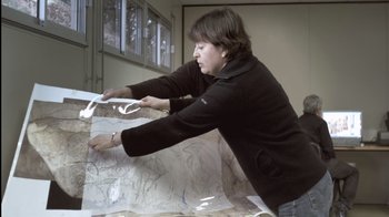 Movie still from “Cave of Forgotten Dreams” (2010), directed by Werner Herzog – A woman in a black jacket is looking at a map; Medium shot, High angle