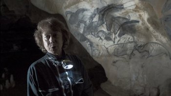 Movie still from “Cave of Forgotten Dreams” (2010), directed by Werner Herzog – An older woman standing in front of a painting of horses; Medium shot, High angle