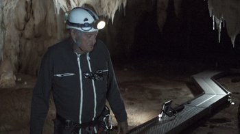 Movie still from “Cave of Forgotten Dreams” (2010), directed by Werner Herzog – A man with a helmet and a flashlight on his head is standing next to a platform; Wide shot, High angle
