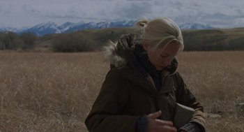 Movie still from “Certain Women” (2016), directed by Kelly Reichardt – A woman in a brown jacket standing in a field looking at her cell phone; Medium shot, Over the shoulder angle