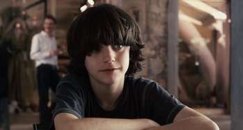 Movie still from “Certified Copy” (2010), directed by Abbas Kiarostami – A young boy with long black hair sitting at a table with his arms crossed; Close Up shot, Over the shoulder angle