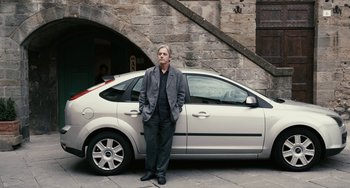 Movie still from “Certified Copy” (2010), directed by Abbas Kiarostami – A man standing in front of a car on a sidewalk; Wide shot, Over the shoulder angle