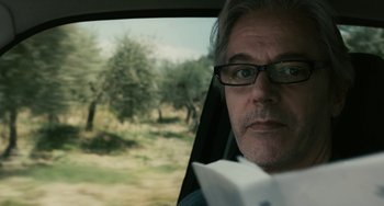 Movie still from “Certified Copy” (2010), directed by Abbas Kiarostami – A man sitting in the passenger seat of a car reading a book; Close Up shot, Low angle