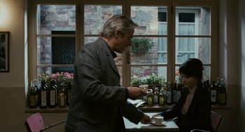 Movie still from “Certified Copy” (2010), directed by Abbas Kiarostami – A man standing in front of a table with a cup of coffee; Medium shot, Over the shoulder angle