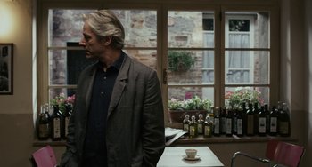 Movie still from “Certified Copy” (2010), directed by Abbas Kiarostami – A man standing in front of a table with a cup of coffee; Medium shot, Over the shoulder angle