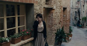 Movie still from “Certified Copy” (2010), directed by Abbas Kiarostami – A woman in a dress is standing in front of a brick building; Wide shot, High angle
